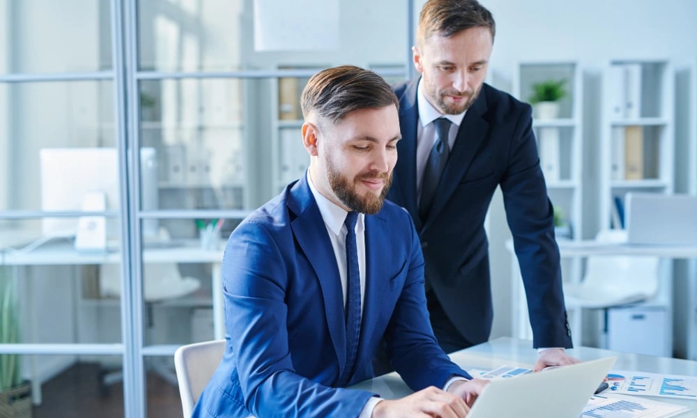 Two brokers in tailored suits reviewing a deal together at a laptop in a bright office