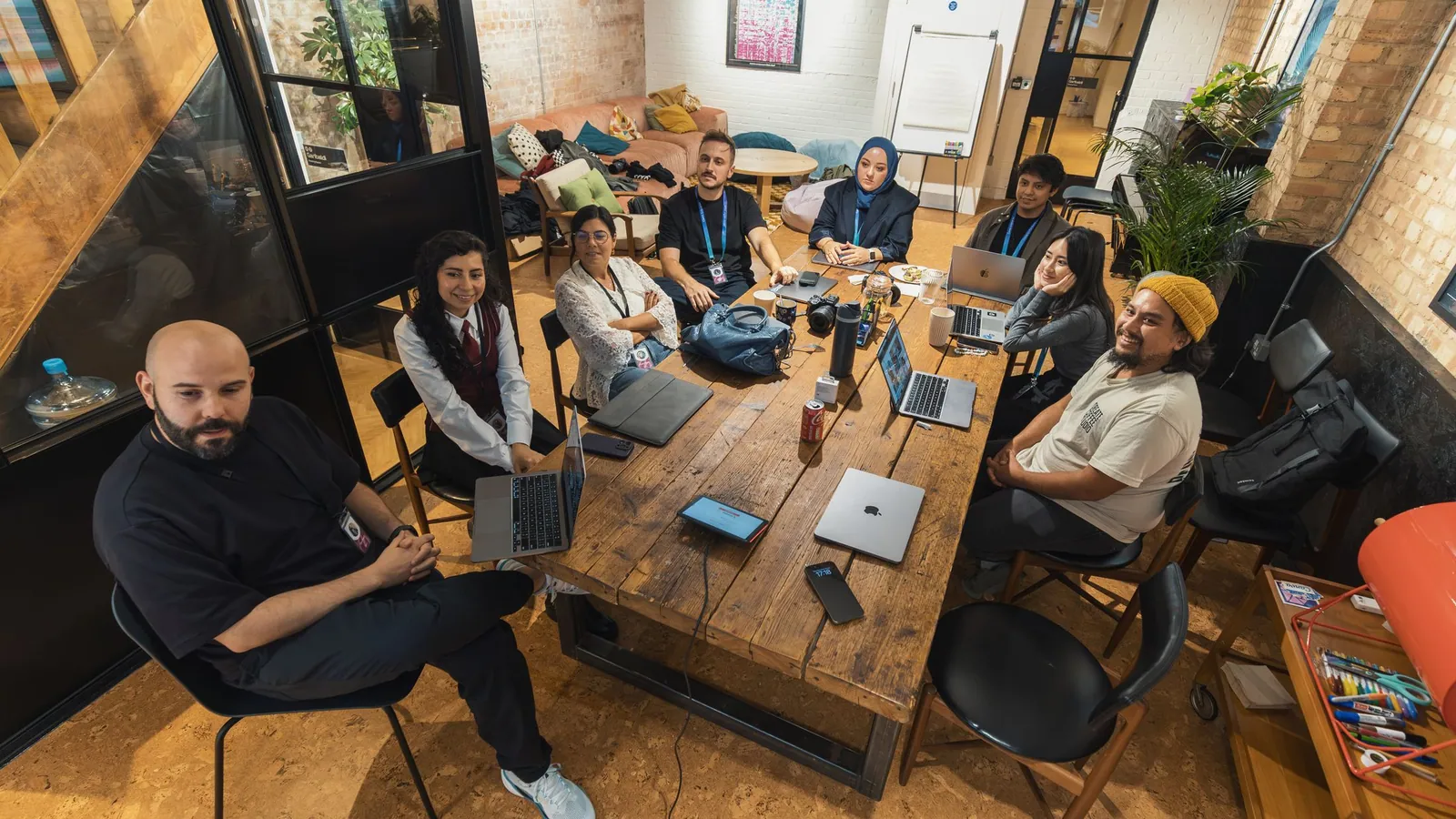 A diverse team gathered around a long wooden table with laptops in a warm, open workspace