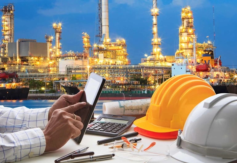 Engineer reviewing a tablet beside hard hats and plans, with an illuminated refinery behind